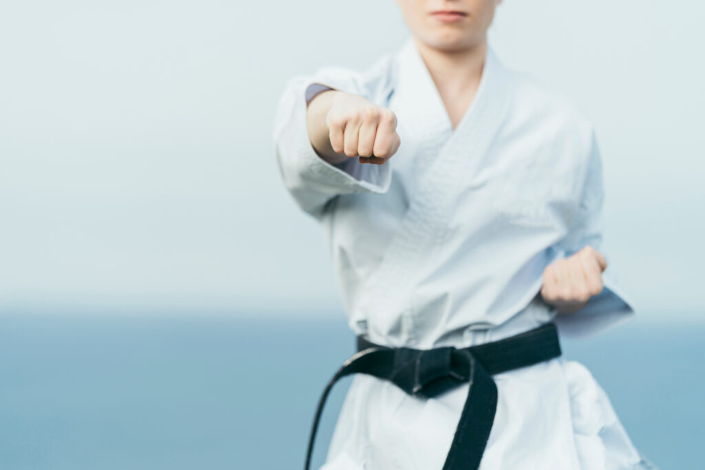 Person in a white martial arts uniform and black belt performs a punch, standing outdoors with a blurred blue background.