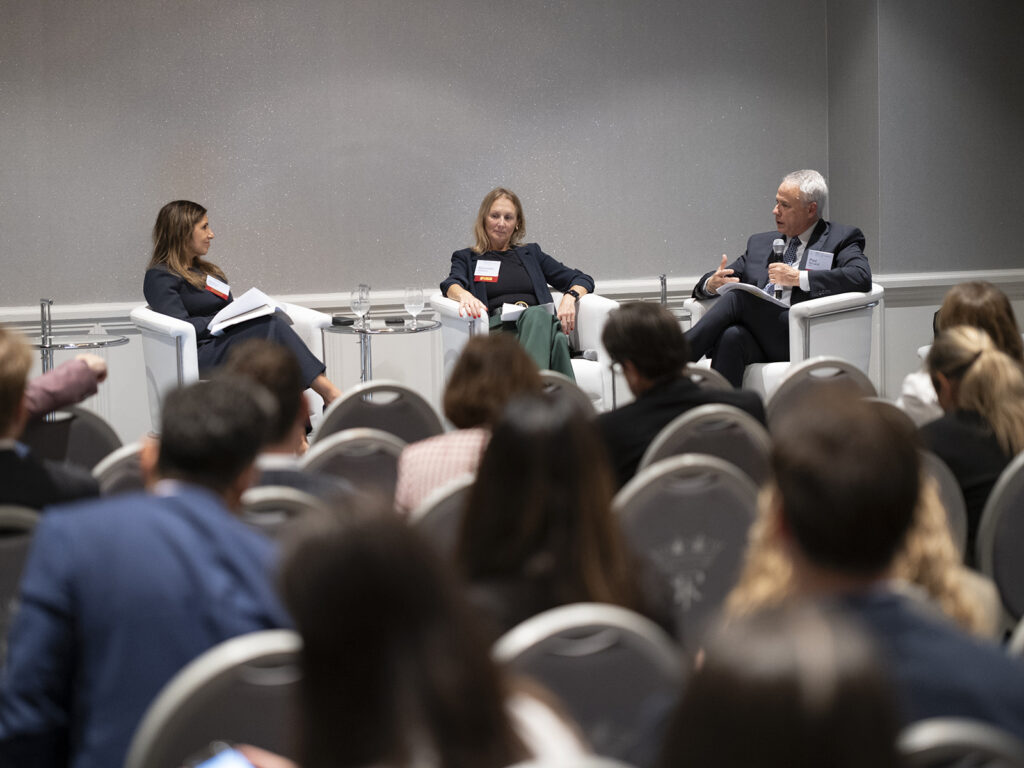 Three panelists sit on stage in white chairs, speaking to an audience during a conference or seminar in a formal setting.