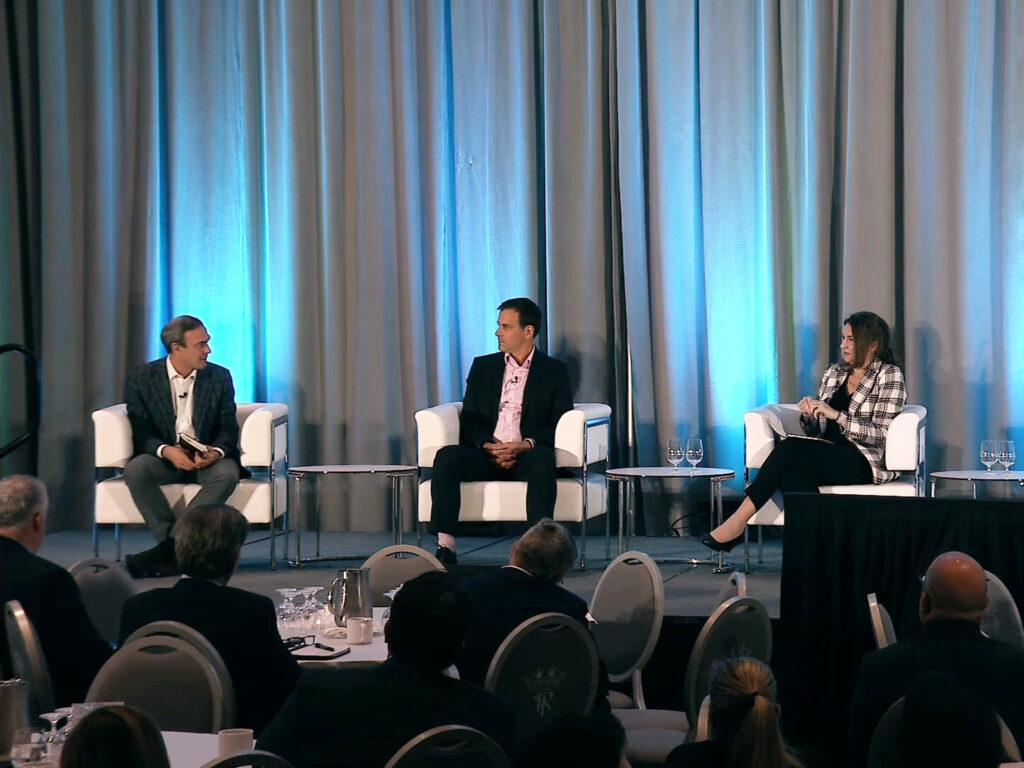 Three people sit on stage in armchairs, engaged in a panel discussion at a conference with an audience seated in front of them.