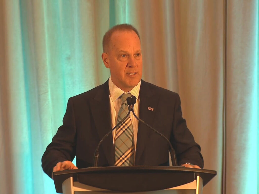 A man in a suit speaks at a podium with two microphones in front of closed curtains.