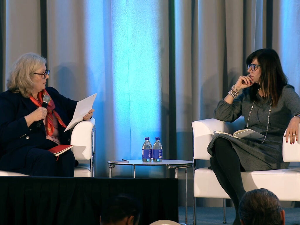 Two women sit on stage in white chairs having a discussion. One holds a microphone and papers, the other listens with a notebook. Two water bottles are on a small table between them.