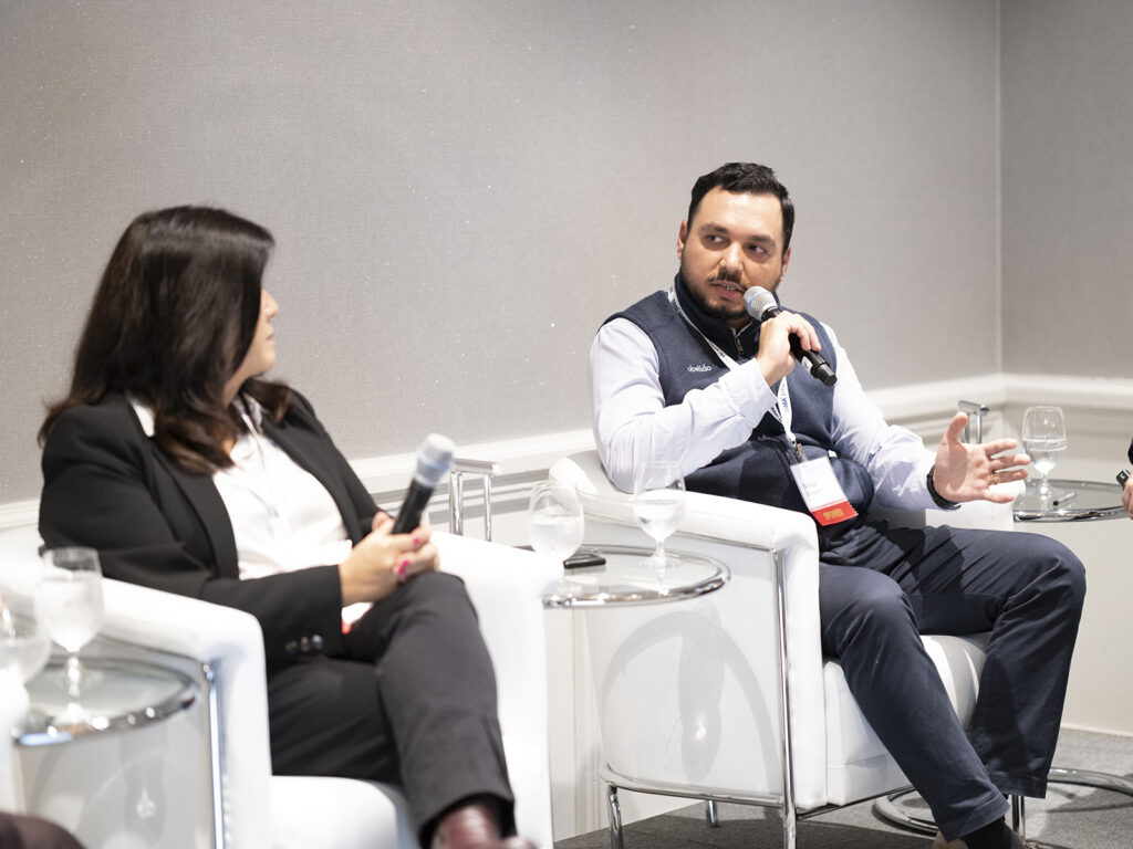 A man speaking into a microphone sits in a white chair next to a woman holding a microphone at a conference.