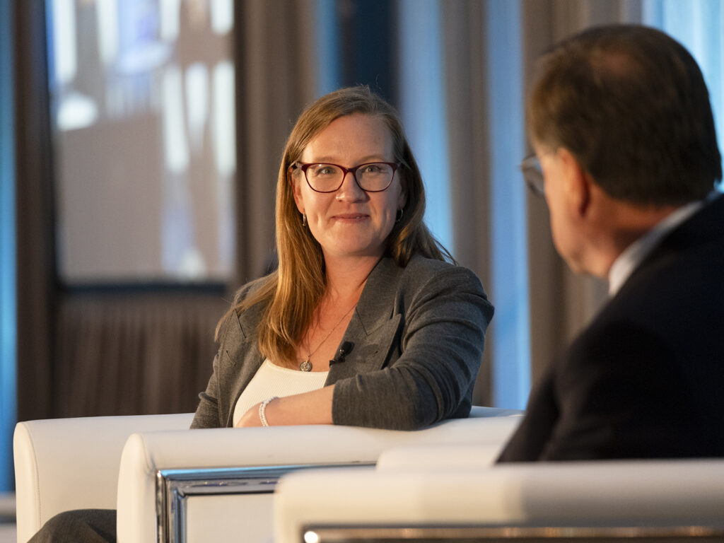 A woman in business attire sits on a white chair, listening and smiling at a man during a professional event or interview.