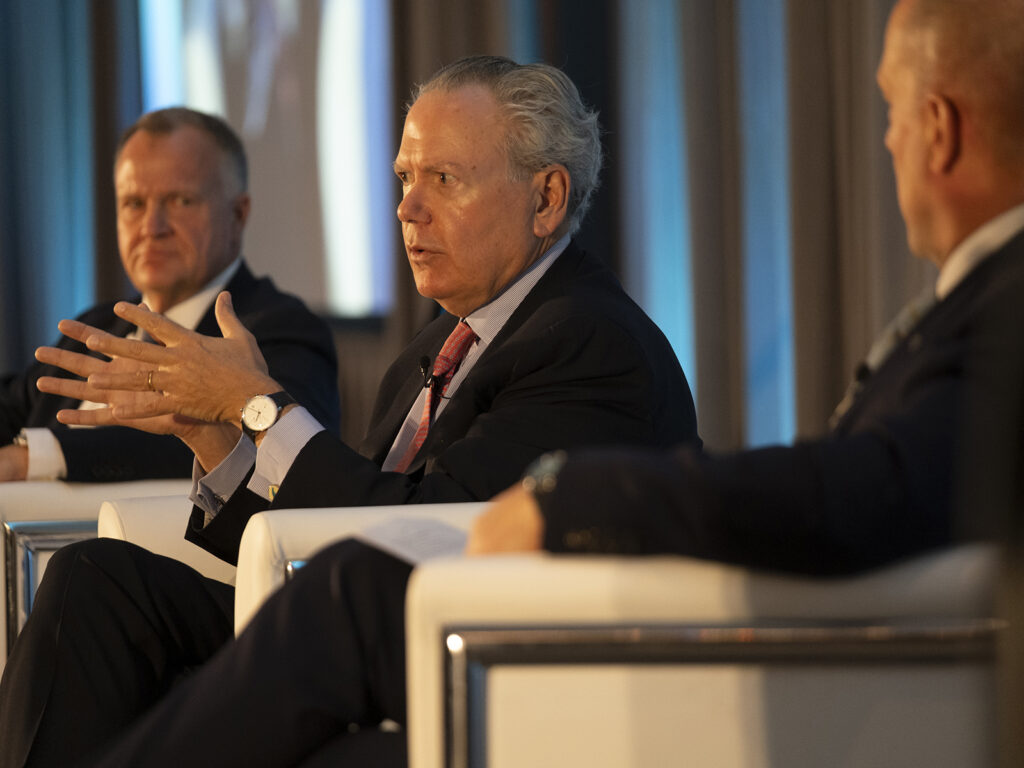 Three men in suits sit on stage in white chairs, one speaking and gesturing with his hands, during a formal panel discussion or conference.