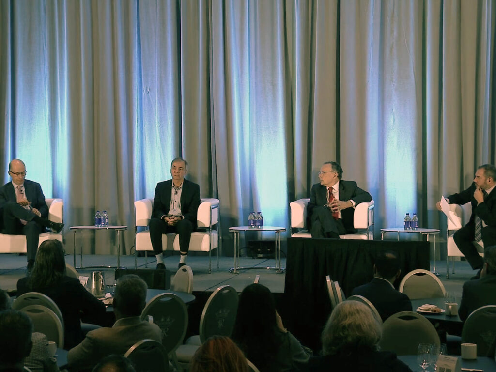 Four men in suits sit on a stage panel in front of an audience, with water bottles on small tables and blue curtains in the background.