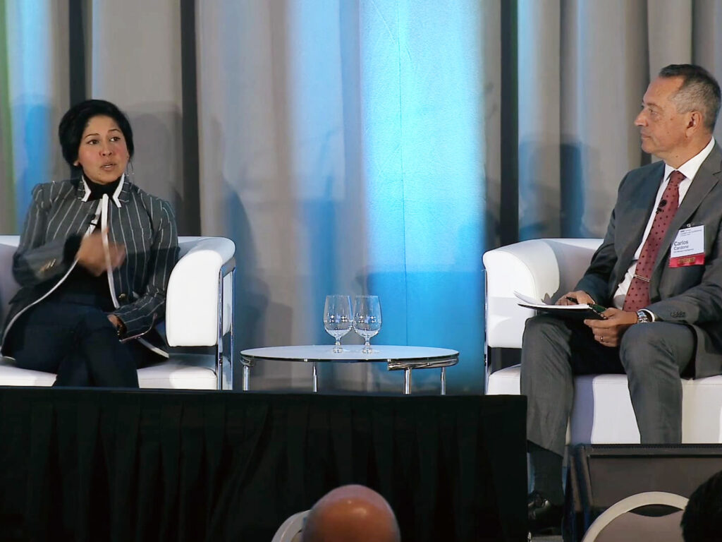 Two people in business attire sit on stage in white chairs, engaged in conversation, with a small table and two glasses between them and a blue-gray curtain backdrop.
