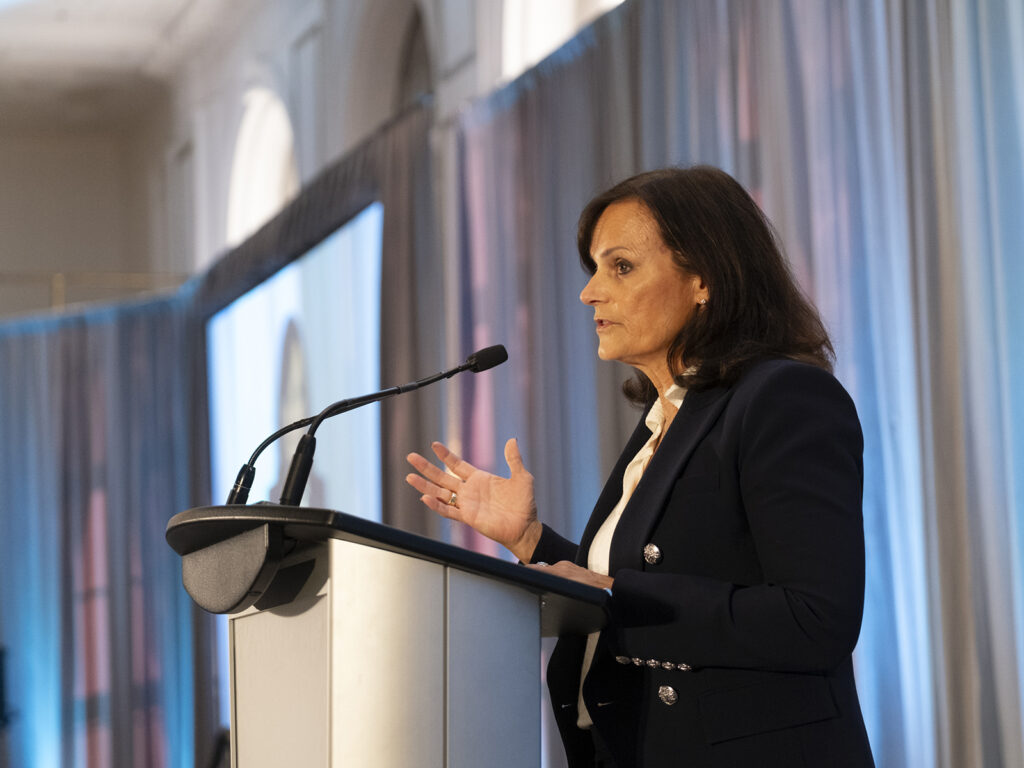 A woman in a dark blazer speaks at a podium with a microphone in front of curtains at an indoor event or conference.
