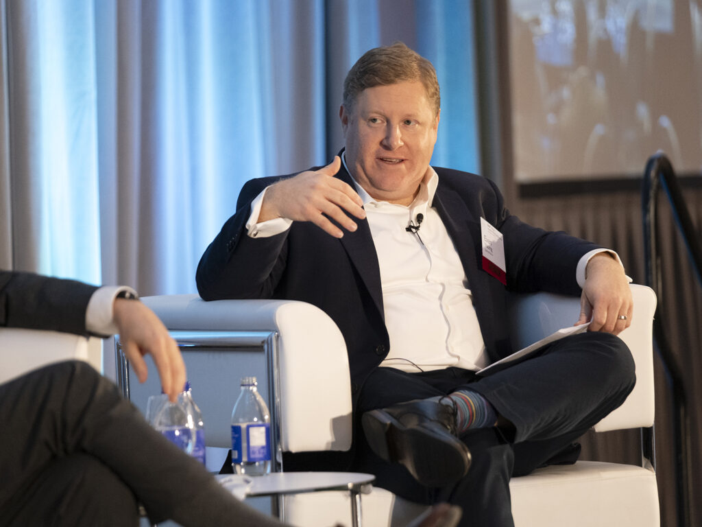 Man in a suit sits on a white chair, speaking and gesturing with his hand during a panel discussion; two water bottles and another person’s arm are visible.