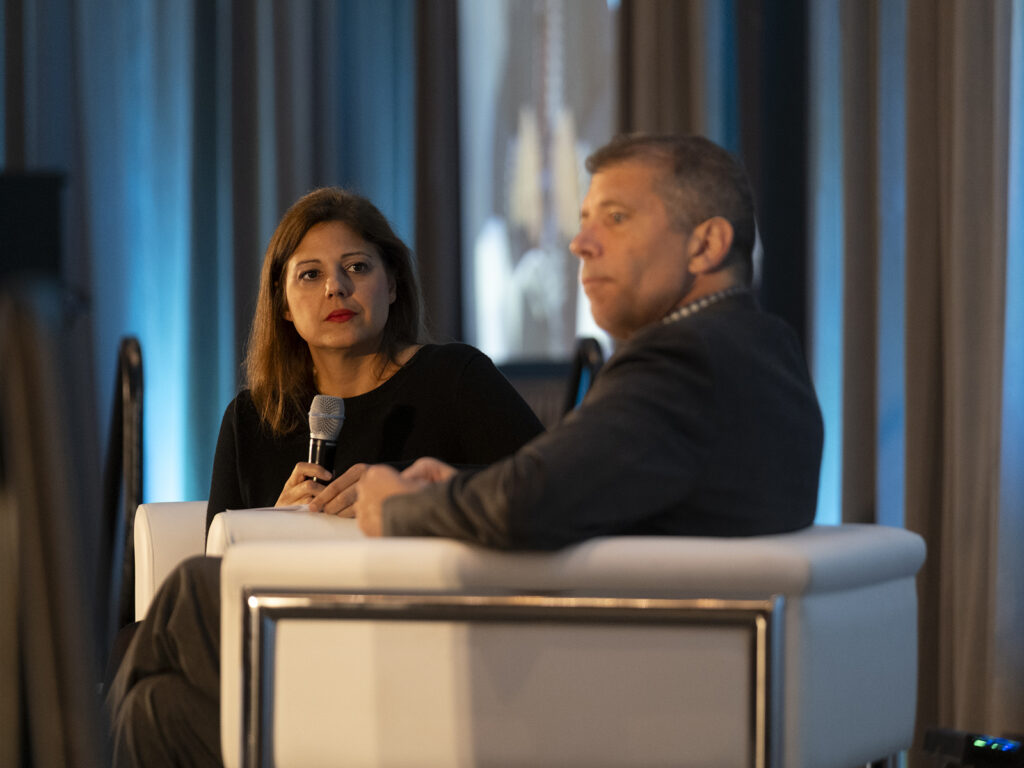 Two people sit on white chairs in conversation; the woman holds a microphone while listening to the man speaking. A blue backdrop with curtains is visible behind them.