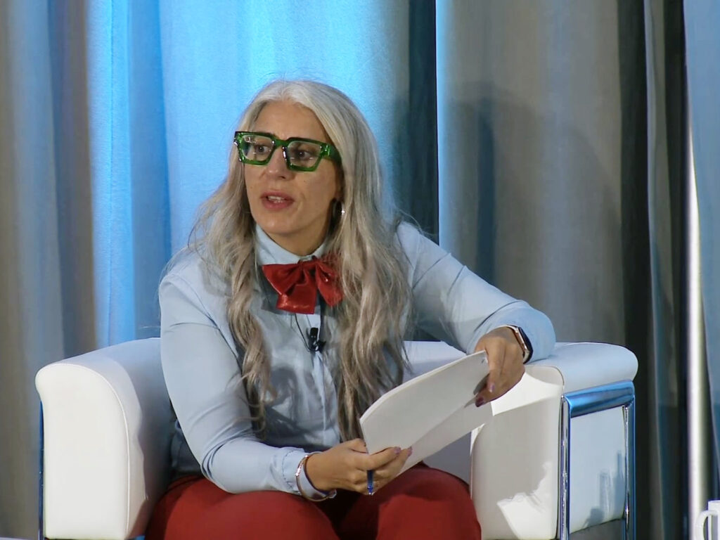 A woman with long gray hair, glasses, and a red bow tie sits in a white chair holding papers, speaking at an event with a blue curtain background.