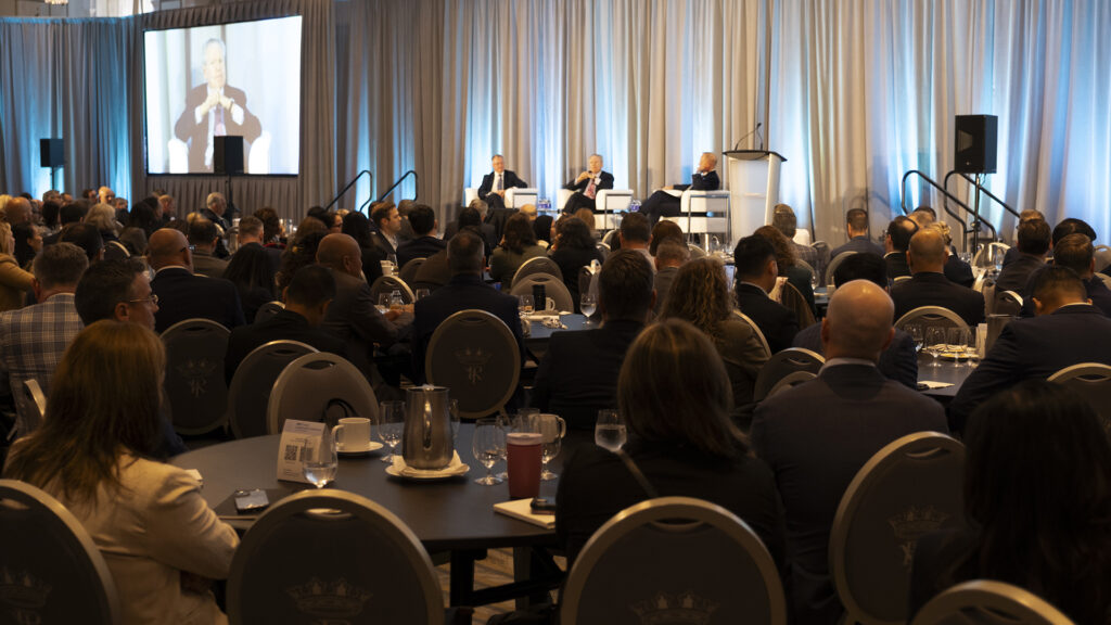 A large audience listens to a panel of speakers on stage at a conference, with one speaker shown on a screen. The room is filled with round tables and attendees facing the stage.