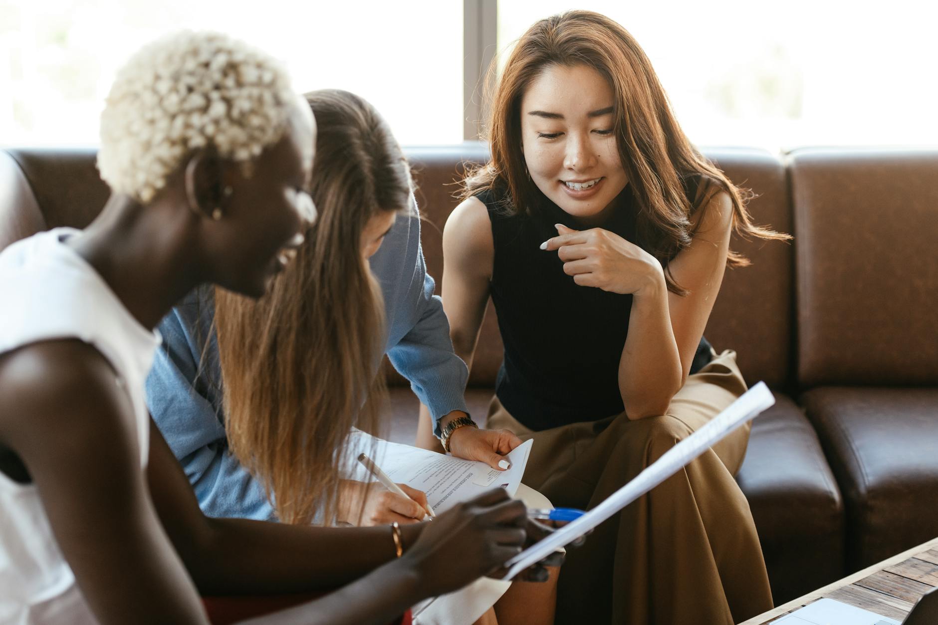 Three women sit on a couch, collaborating over documents and taking notes, with one woman pointing at the papers.