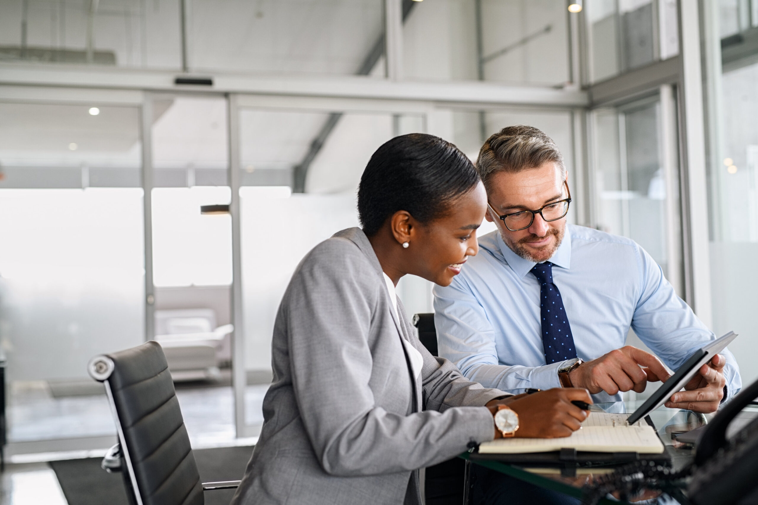 Two business professionals sitting at a desk in an office, discussing information on a tablet while taking notes in a notebook.