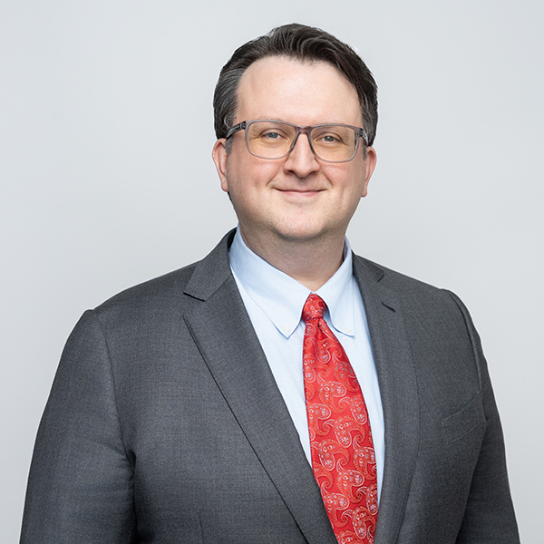 A man in a gray suit, light blue shirt, and red paisley tie stands against a plain light background, smiling at the camera.