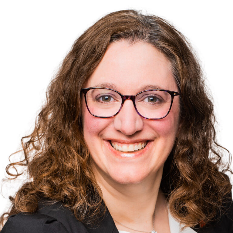 Femme souriante aux longs cheveux bruns bouclés, portant des lunettes, un blazer noir et un haut blanc, sur un fond blanc uni.