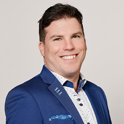 Smiling man with short dark hair wearing a blue blazer and white patterned shirt, standing against a plain light background.