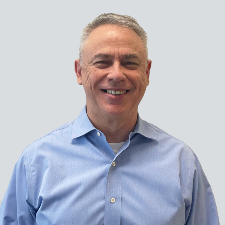 A middle-aged man with short gray hair, wearing a light blue button-up shirt, smiles while standing against a plain light gray background.