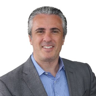 Un homme aux cheveux gris courts, vêtu d'une veste de costume grise et d'une chemise bleue, souriant, posant devant un fond blanc uni.
