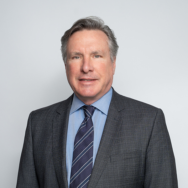 A middle-aged man with gray hair wearing a dark suit, blue shirt, and striped tie, standing against a plain light gray background.