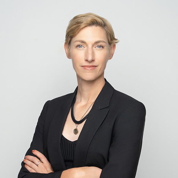 Person with short blond hair in a black blazer and necklace stands with arms crossed against a plain light background, looking directly at the camera with a neutral expression.