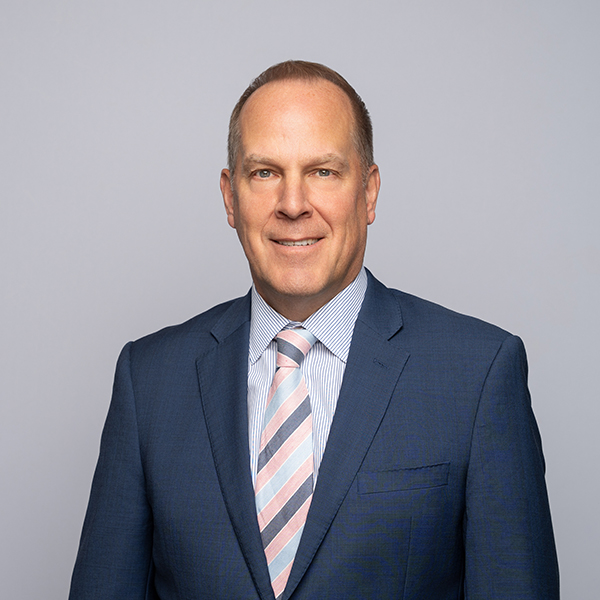 A middle-aged man in a blue suit, light blue striped shirt, and pink striped tie stands in front of a plain light gray background, facing the camera and smiling slightly.