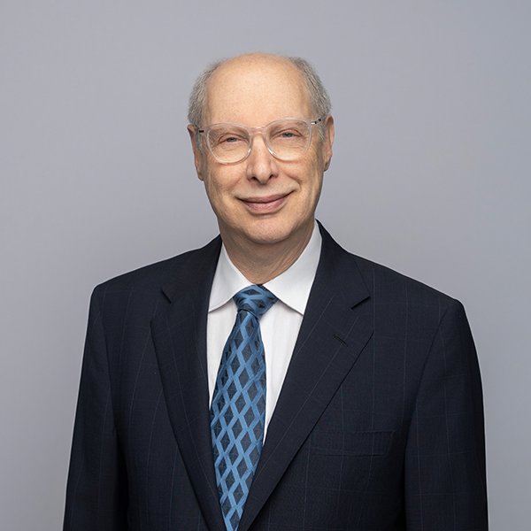 Bald man with glasses in a dark suit, white shirt, and blue patterned tie, posing against a plain light gray background.