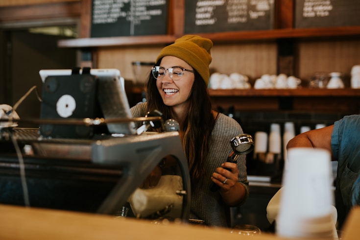 A barista wearing a beanie and glasses smiles while working at a coffee shop counter, operating the espresso machine.