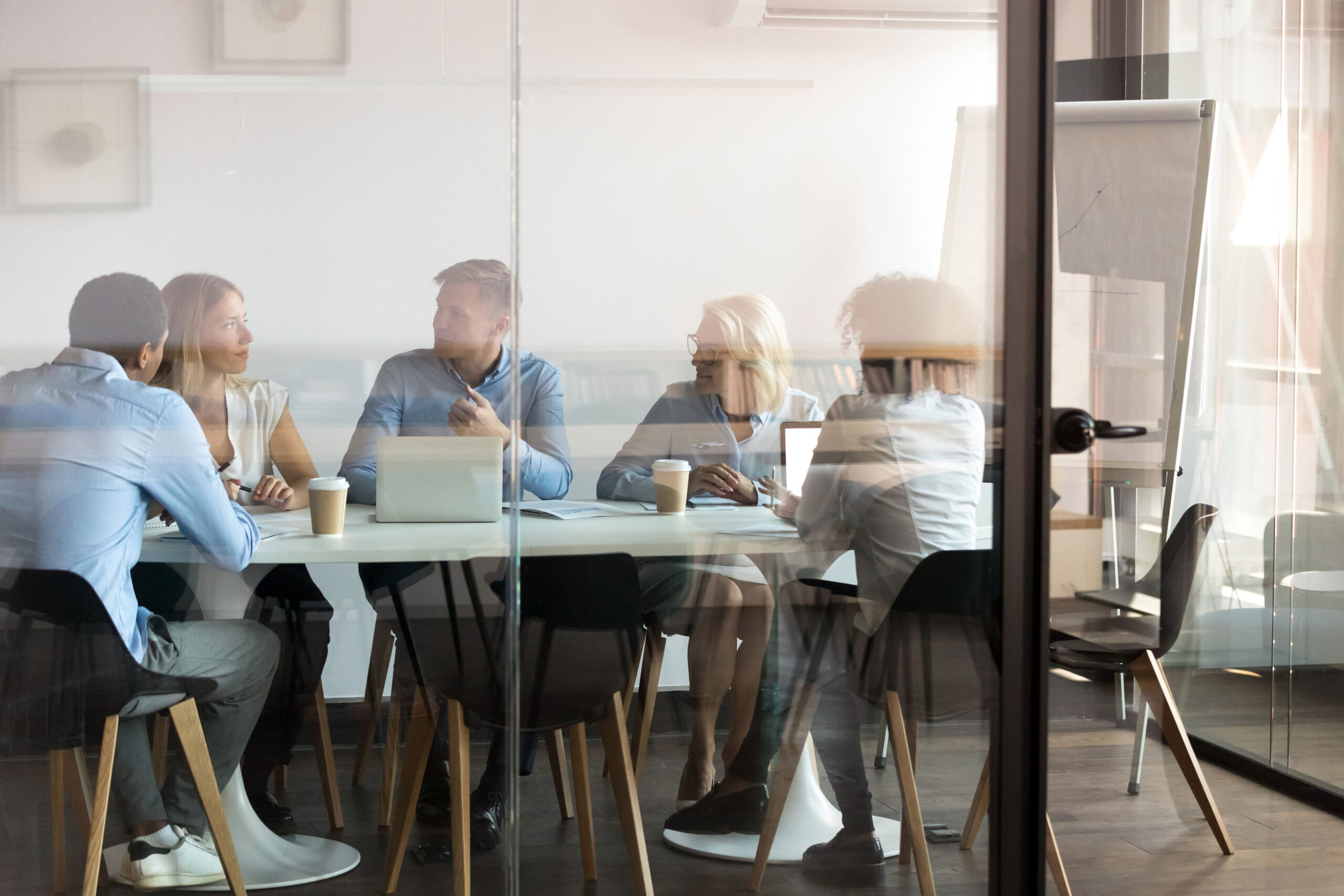 Five people sit around a conference table in a glass-walled meeting room, talking and using laptops, with a flip chart and coffee cups visible.