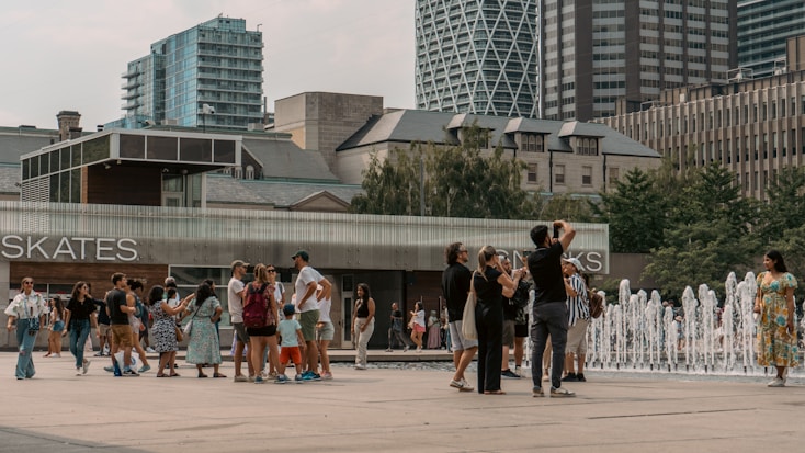 People gather around a fountain in an urban plaza, some taking photos, with modern and older buildings in the background.