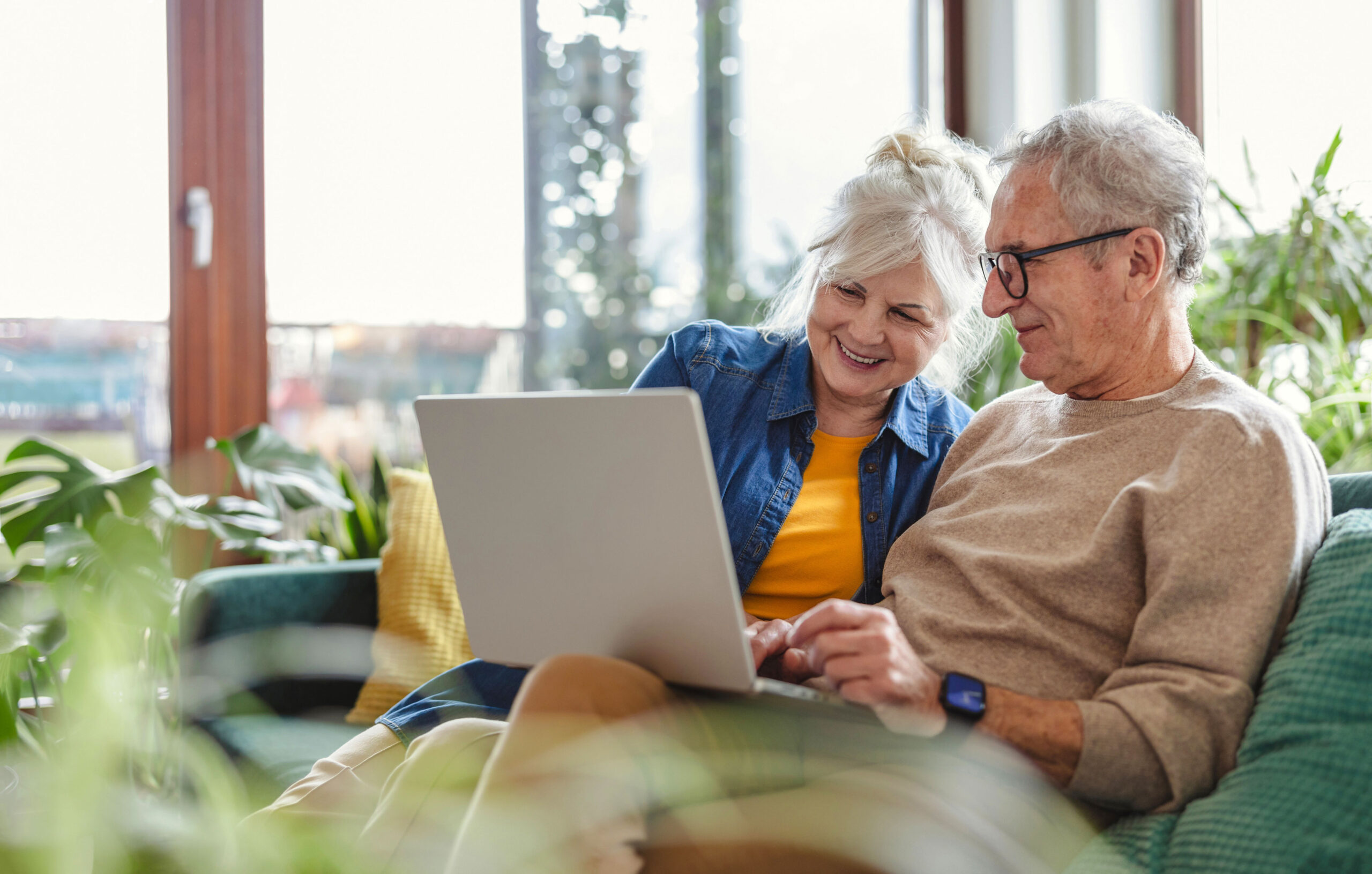 An older couple sits on a couch surrounded by plants, looking at a laptop together and smiling in a brightly lit living room.