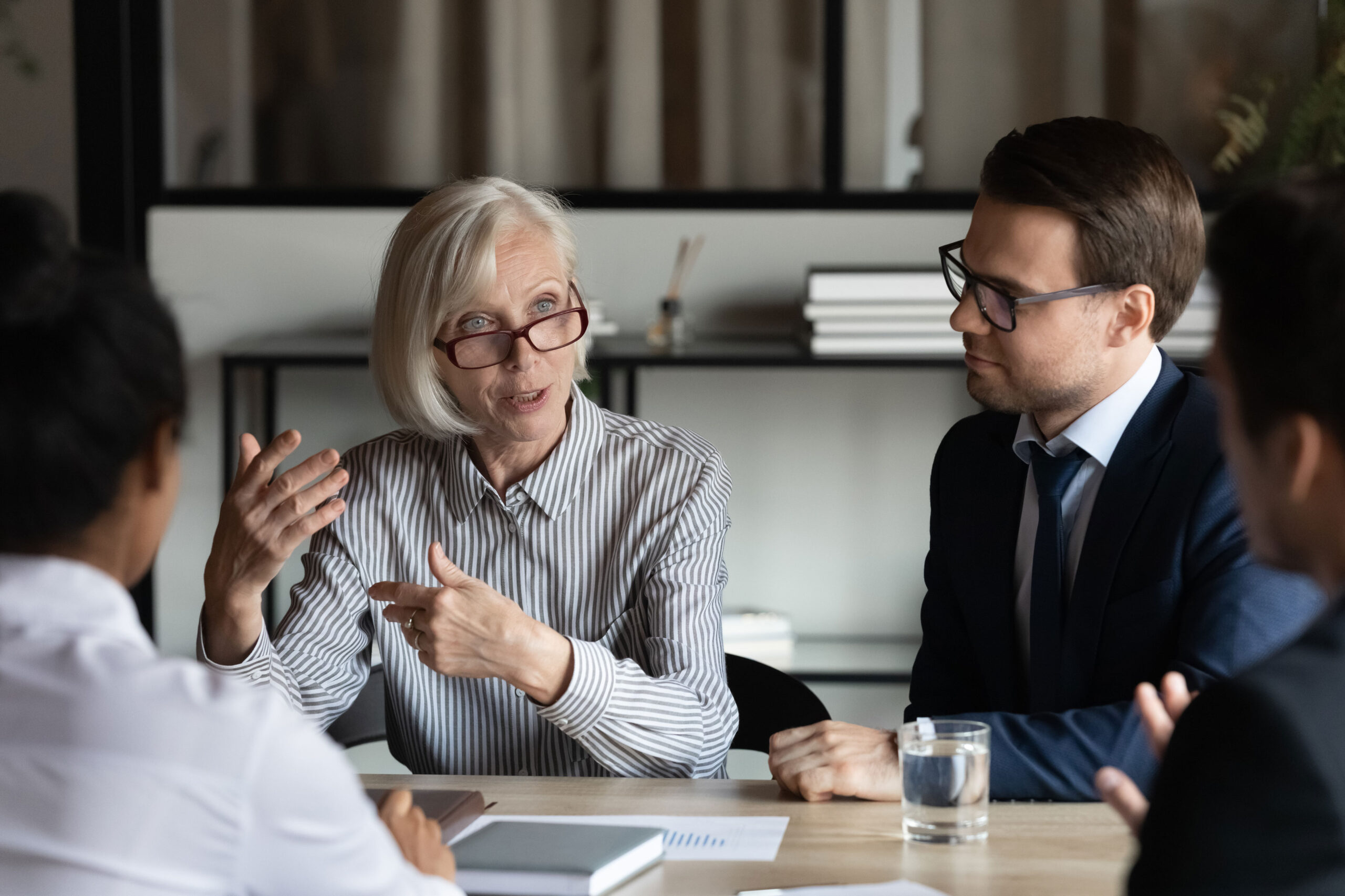 An older woman in glasses speaks to three colleagues at a table during a business meeting, with papers and a glass of water visible.
