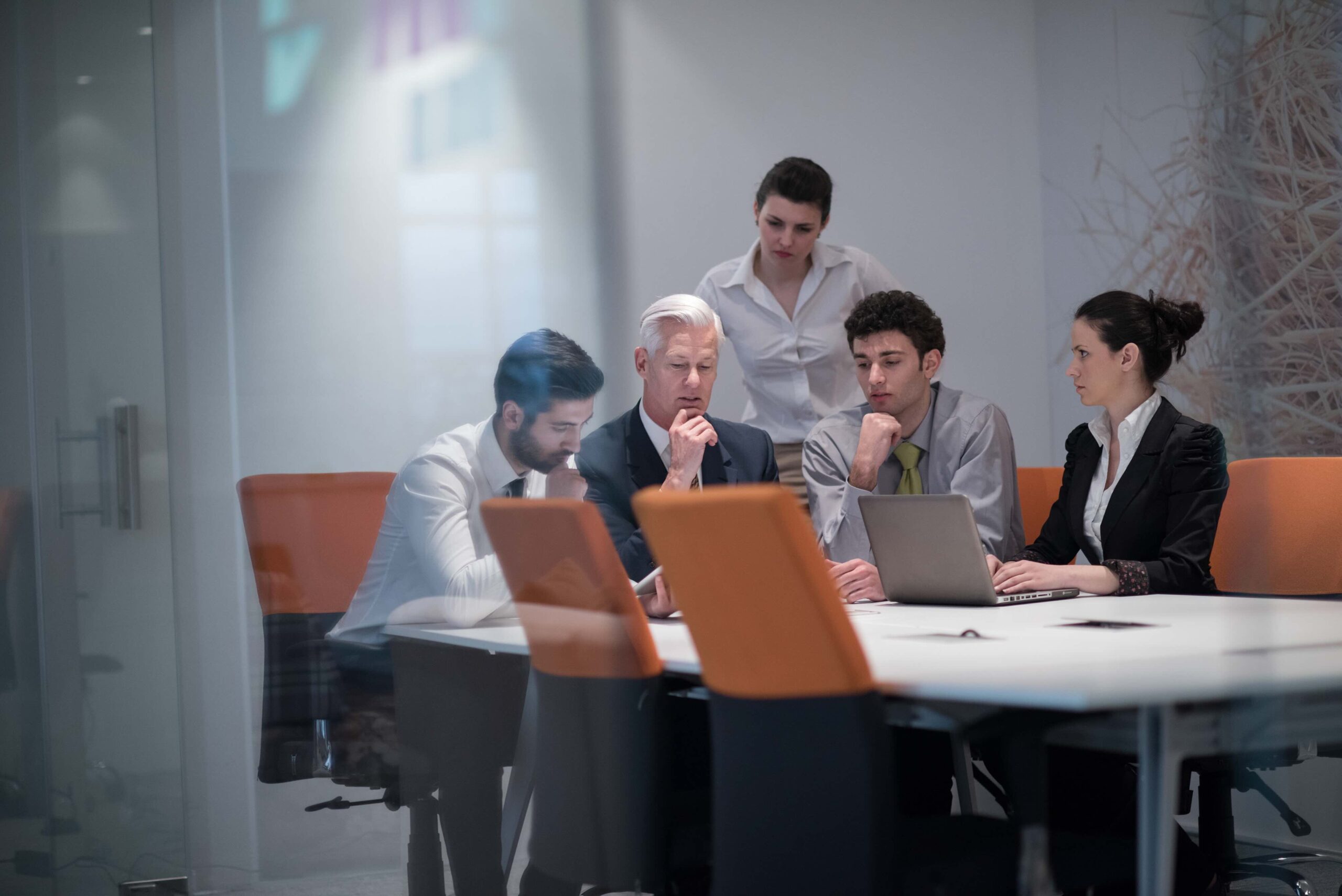 Five people sit and stand around a conference table in an office, looking intently at a laptop and documents during a meeting.
