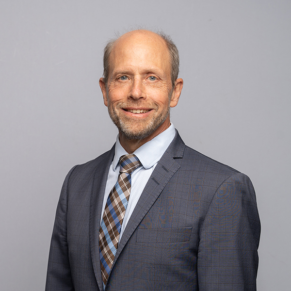 A man with a short beard and receding hairline is wearing a gray suit, blue shirt, and plaid tie, standing against a plain light gray background.