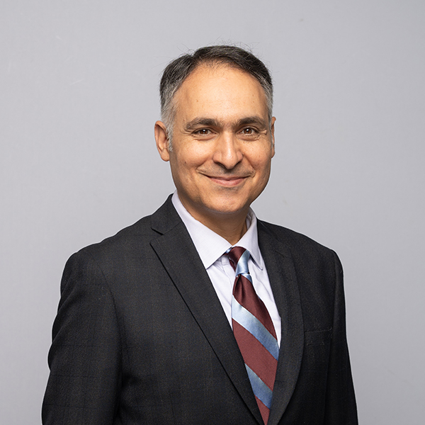 Man in a dark suit, striped tie, and light shirt standing against a plain gray background, smiling at the camera.