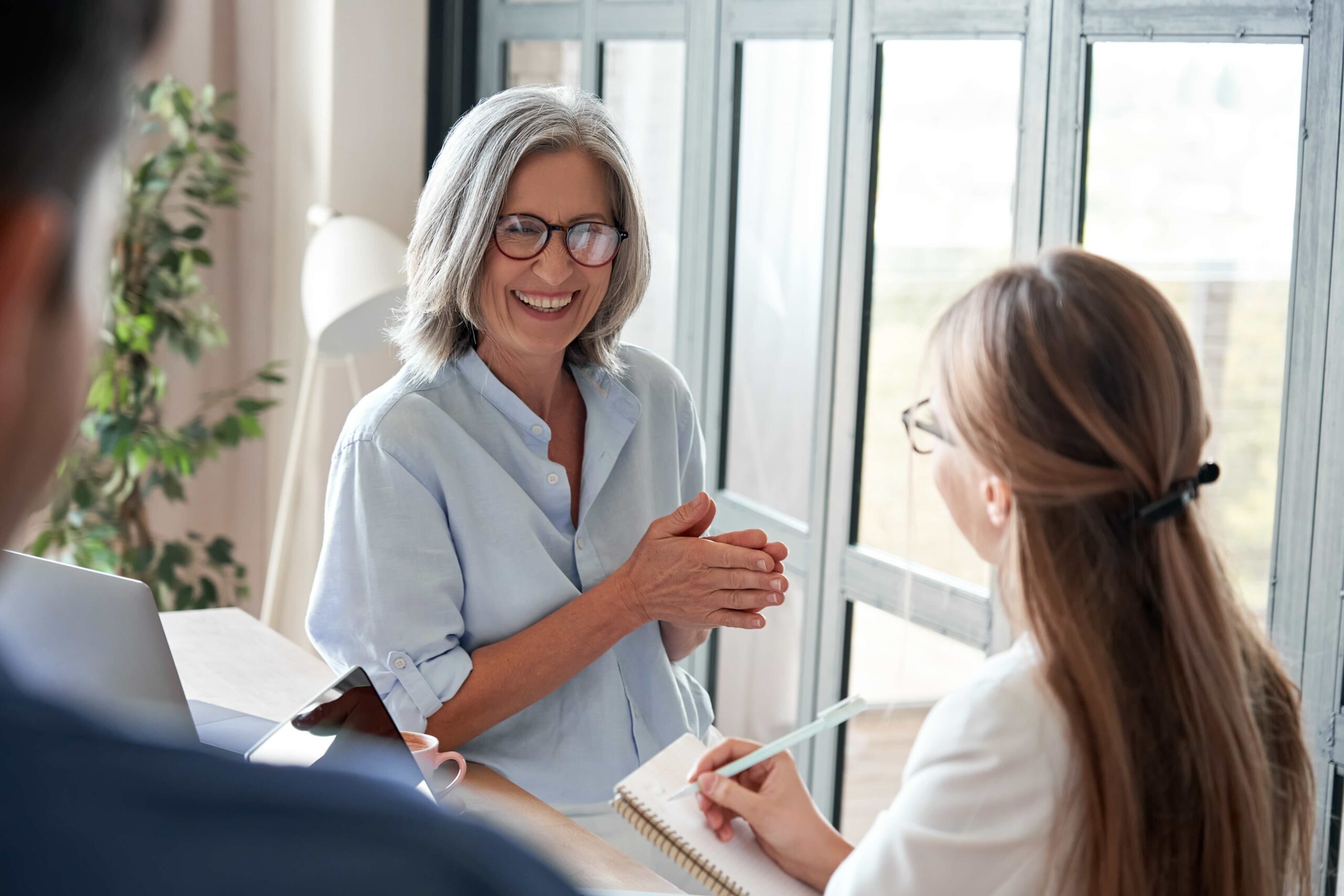 Smiling older woman in glasses talks to two colleagues at a desk. One person writes in a notebook. Large windows and a plant are visible in the background.