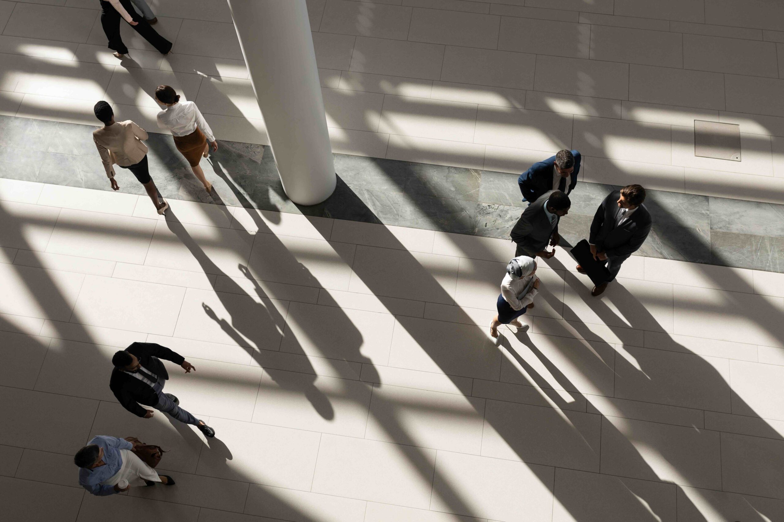 People walk and stand in small groups on a tiled floor, casting long shadows from sunlight streaming through large windows.