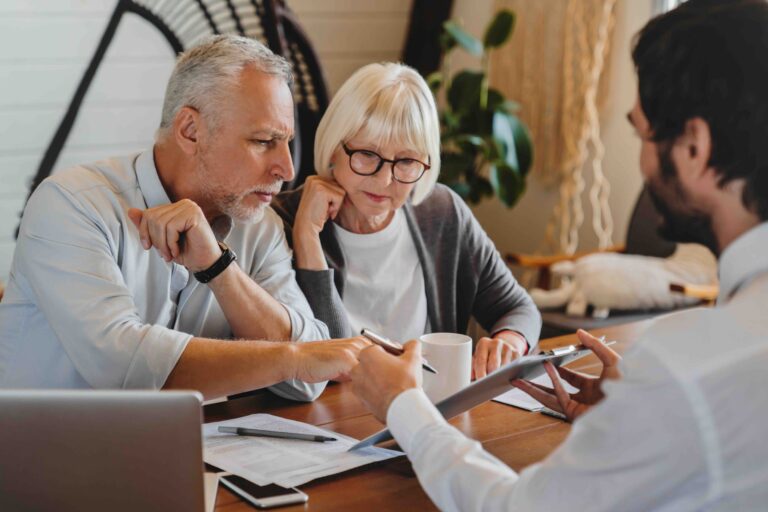 Two older adults sit at a table with paperwork, listening to a man who is showing them information on a tablet.