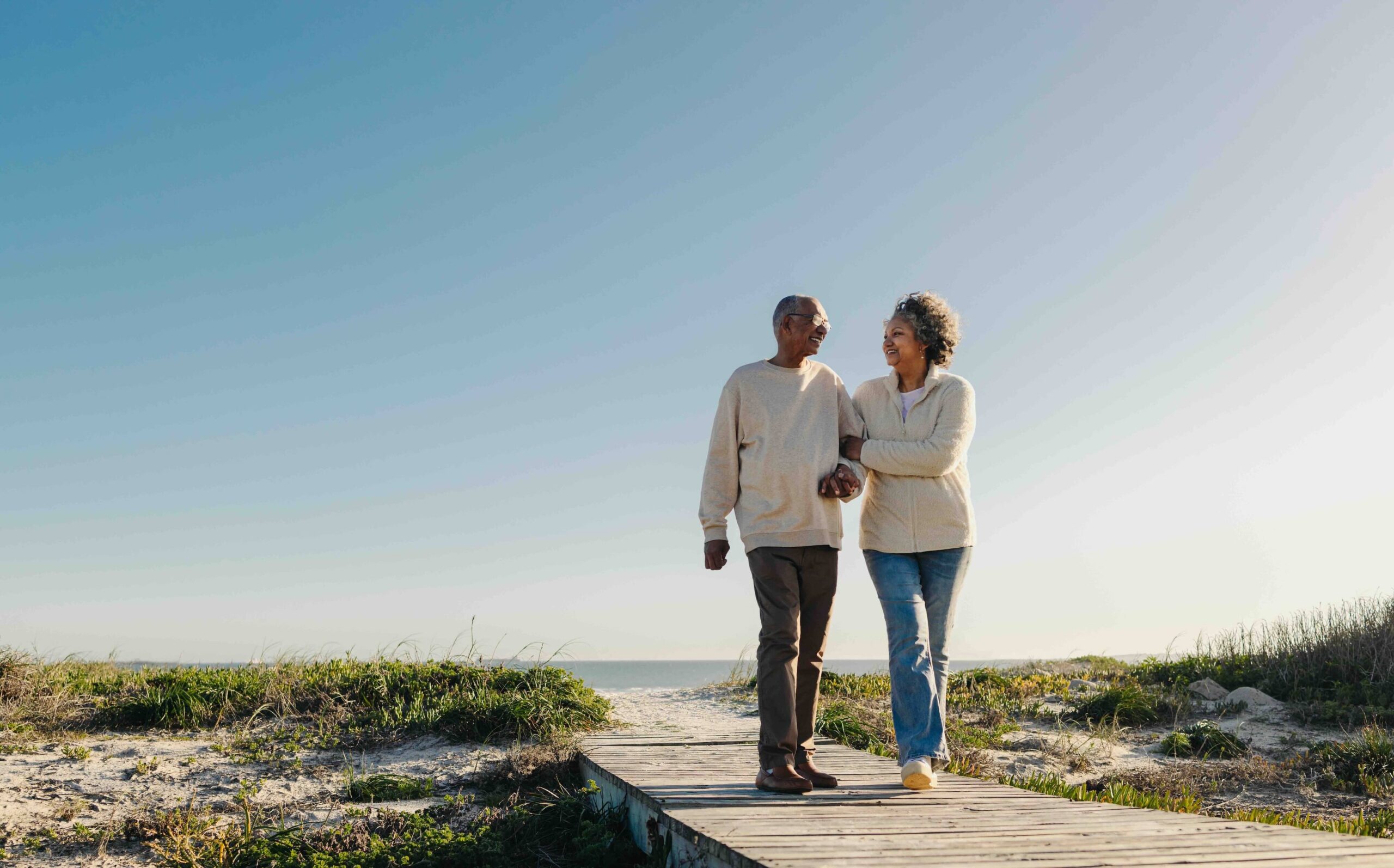 An older couple walks arm in arm on a wooden pathway at the beach, surrounded by sand and greenery, under a clear blue sky.