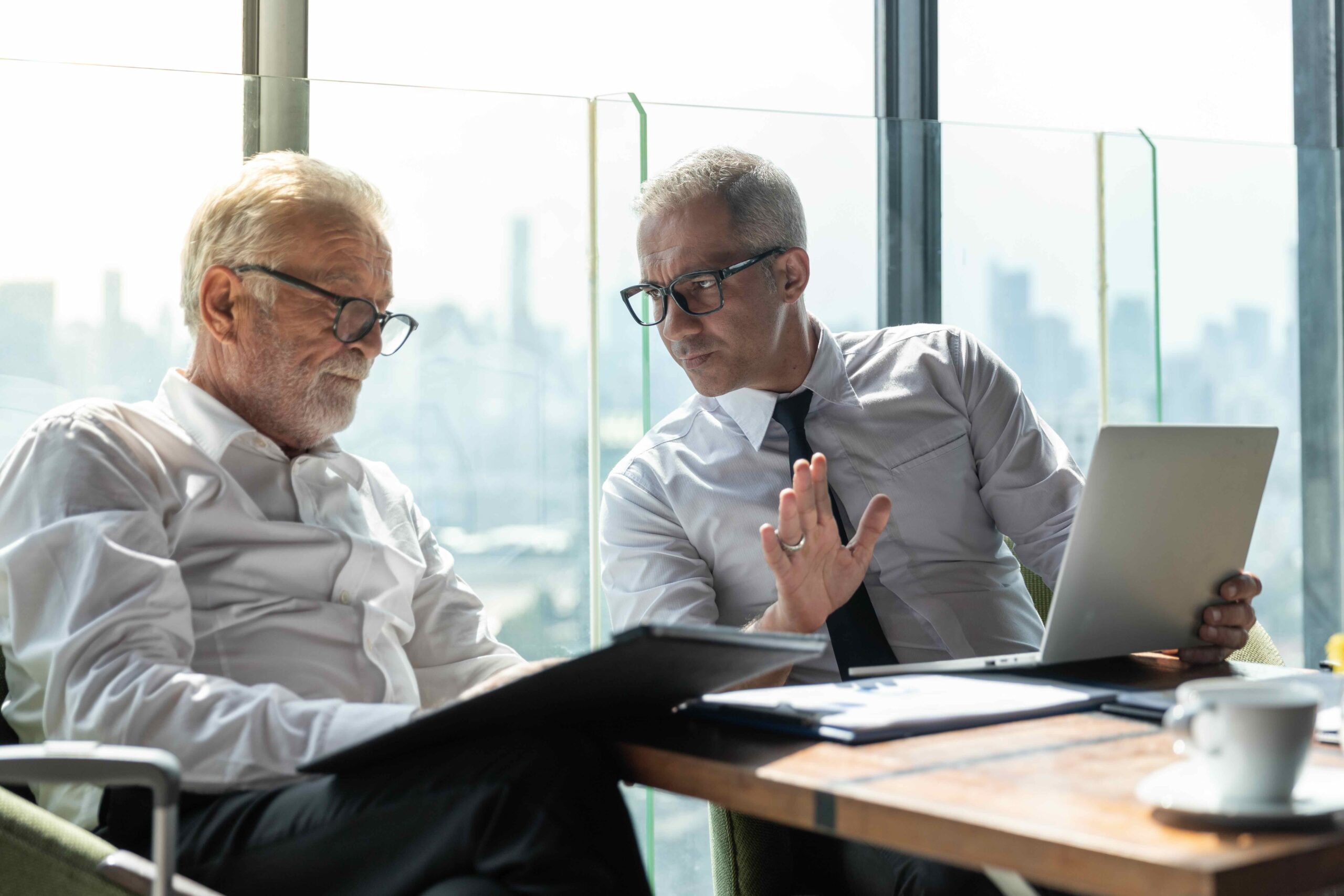 Two men in business attire sit at a table with documents and a laptop, engaged in a serious discussion near large windows overlooking a cityscape.