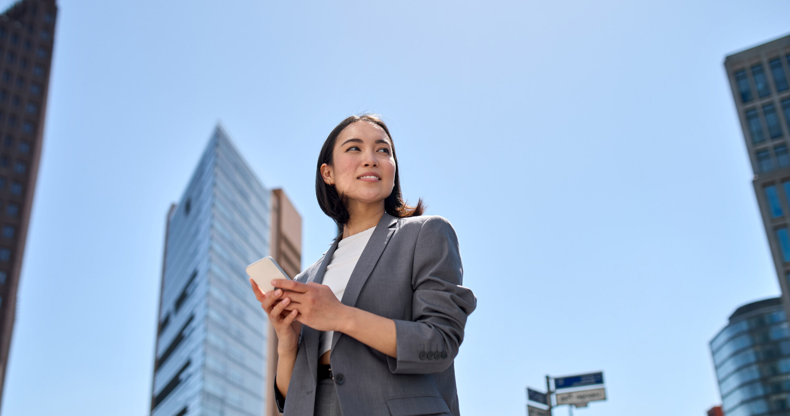 A woman in a gray blazer holds a smartphone and stands outdoors in a city with modern tall buildings under a clear blue sky.