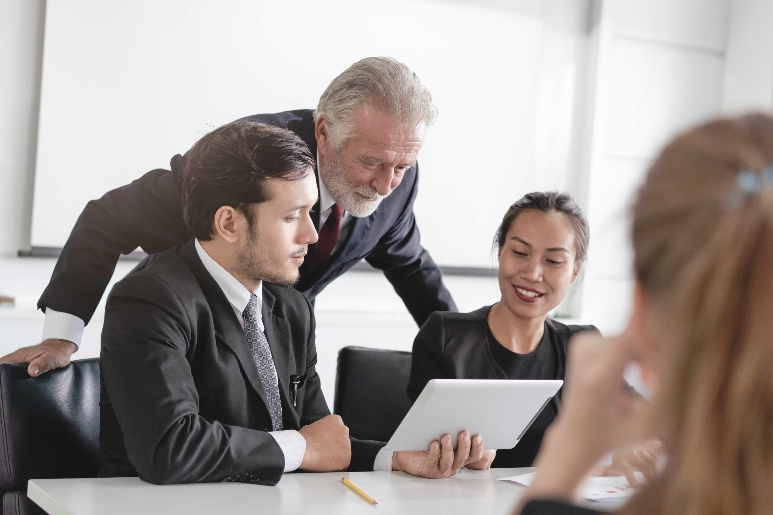 Three people in business attire sit at a table, looking at a tablet together, while another person is partially visible in the foreground.