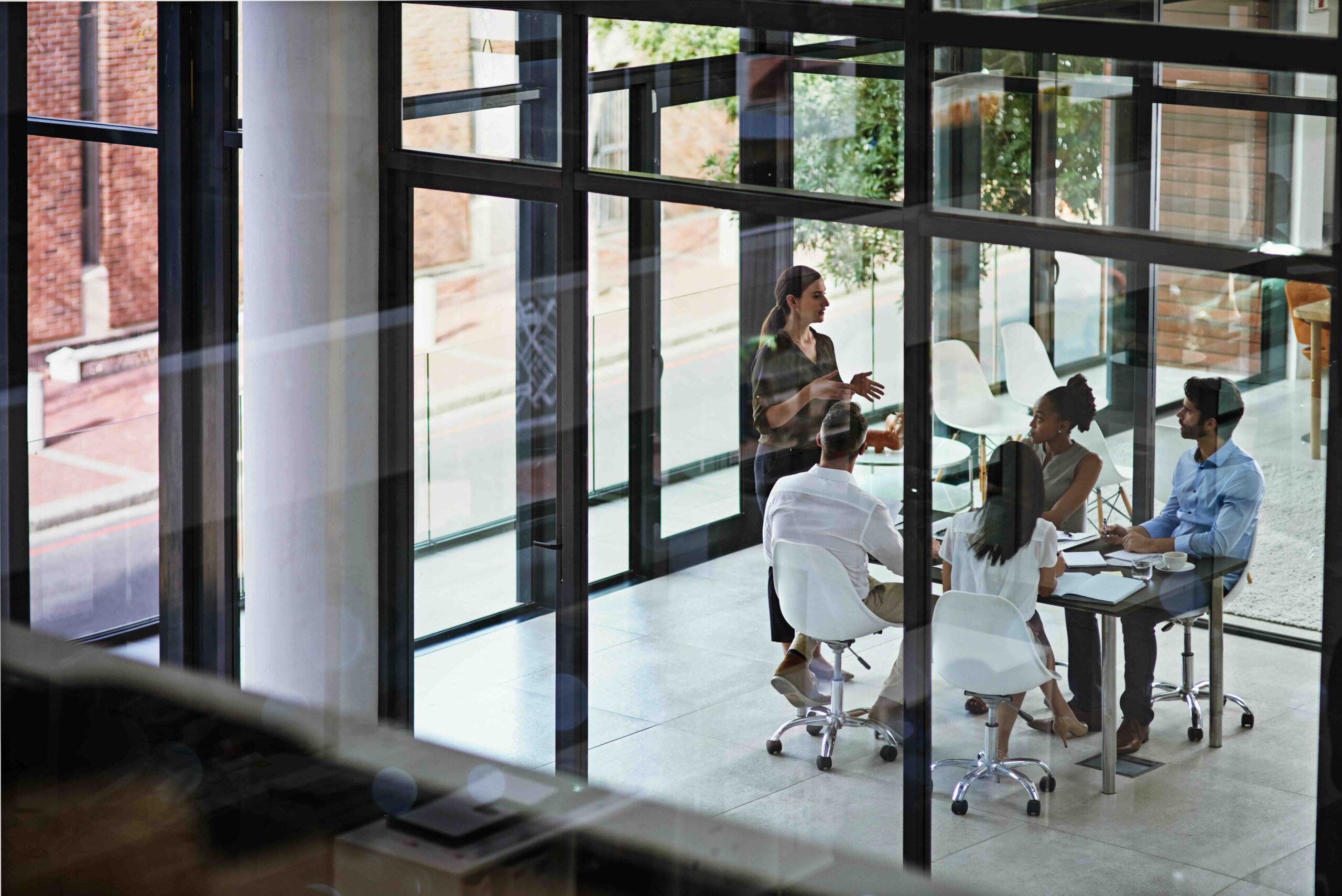 Four people seated at a conference table in a glass-walled office listen as one person stands and speaks, with city buildings visible outside.