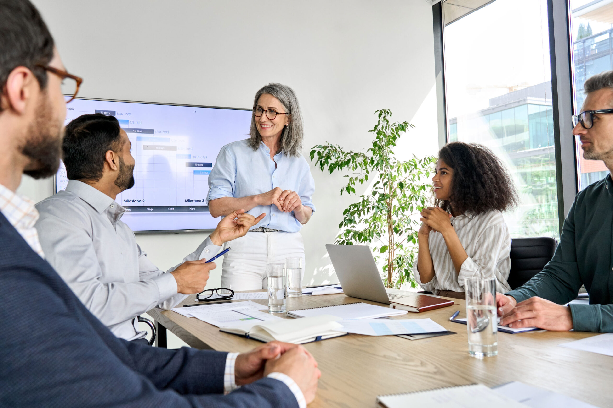 Five people in business attire sit around a conference table, with one woman standing and smiling while leading a meeting; laptops, papers, and glasses of water are on the table.