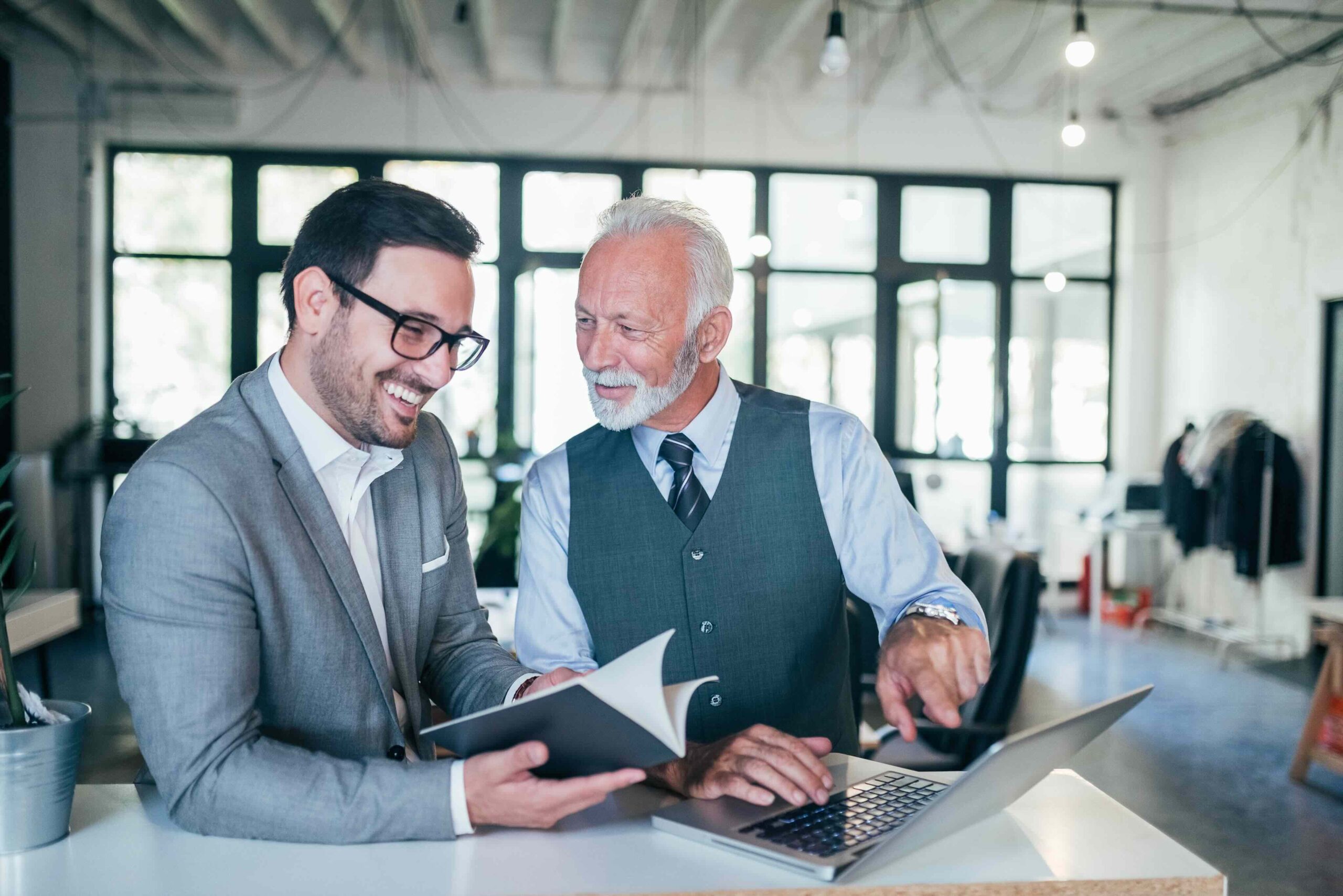 Two men in business attire discuss work at a desk with a laptop and notebook in a modern office setting, both appearing engaged and smiling.