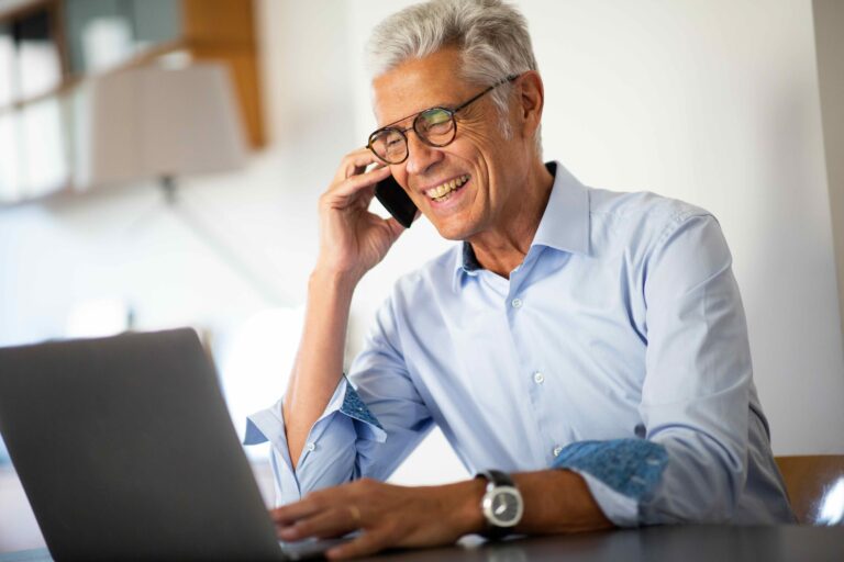 Older man wearing glasses and a light blue shirt sits at a desk, smiling while talking on a smartphone and working on a laptop.