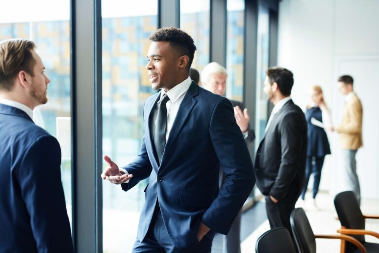 Two men in business suits have a conversation in an office with large windows, while other professionals talk in the background.