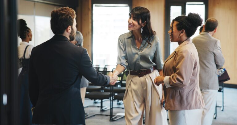 A woman in business attire shakes hands with a man in a suit while another woman stands beside them in a conference room setting.