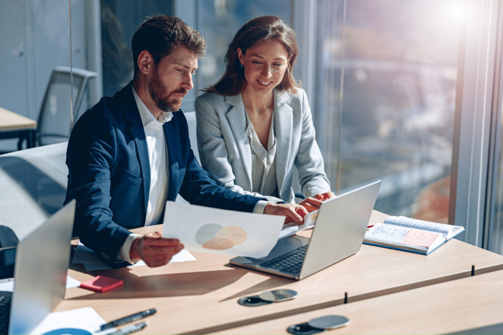 Deux professionnels sont assis à un bureau ; ils consultent un graphique imprimé et travaillent sur un ordinateur portable dans un bureau baigné de soleil, avec des cahiers et des documents posés sur la table.