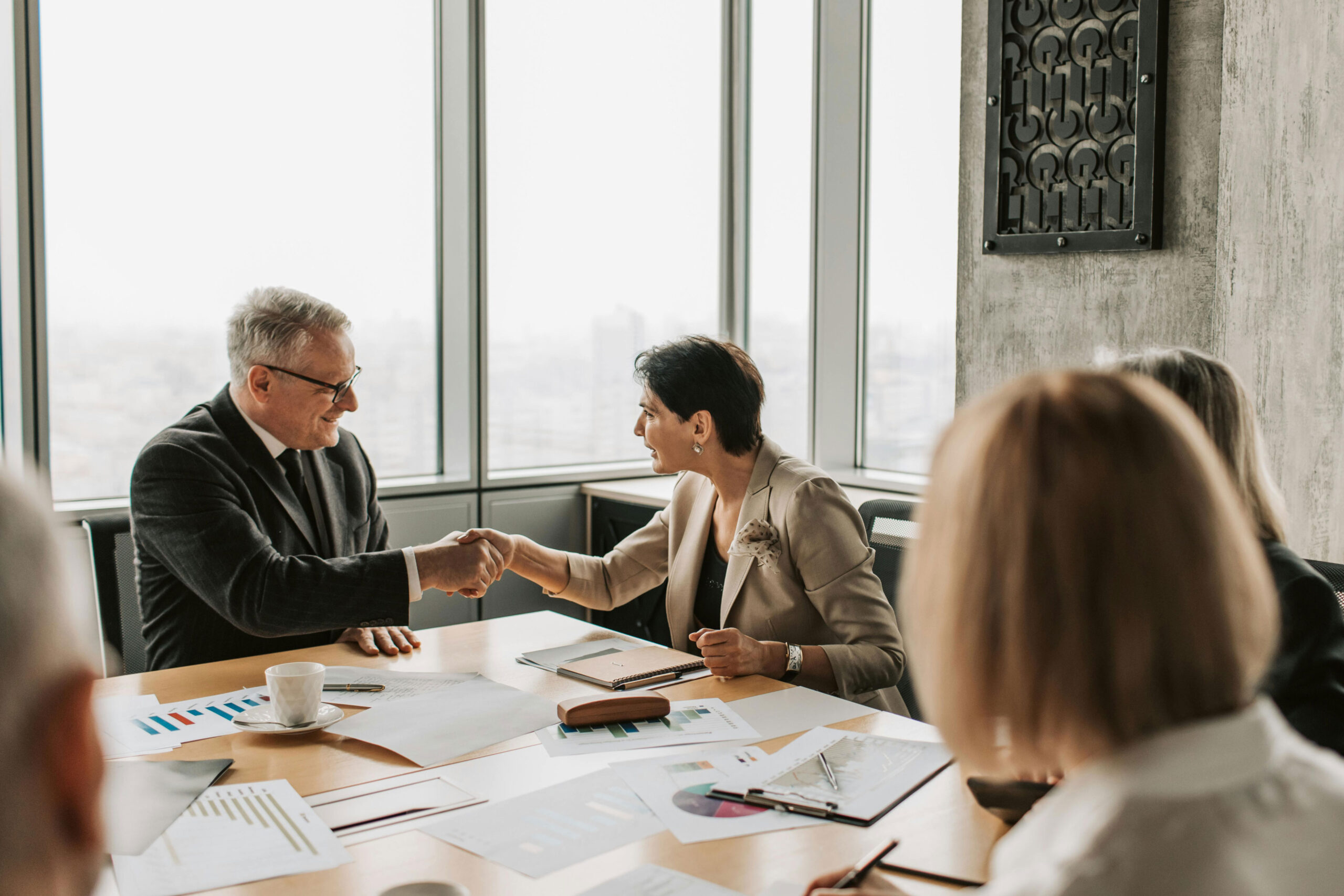 Two businesspeople shake hands across a table in a modern office meeting room, with documents, charts, and coffee cups visible on the table.
