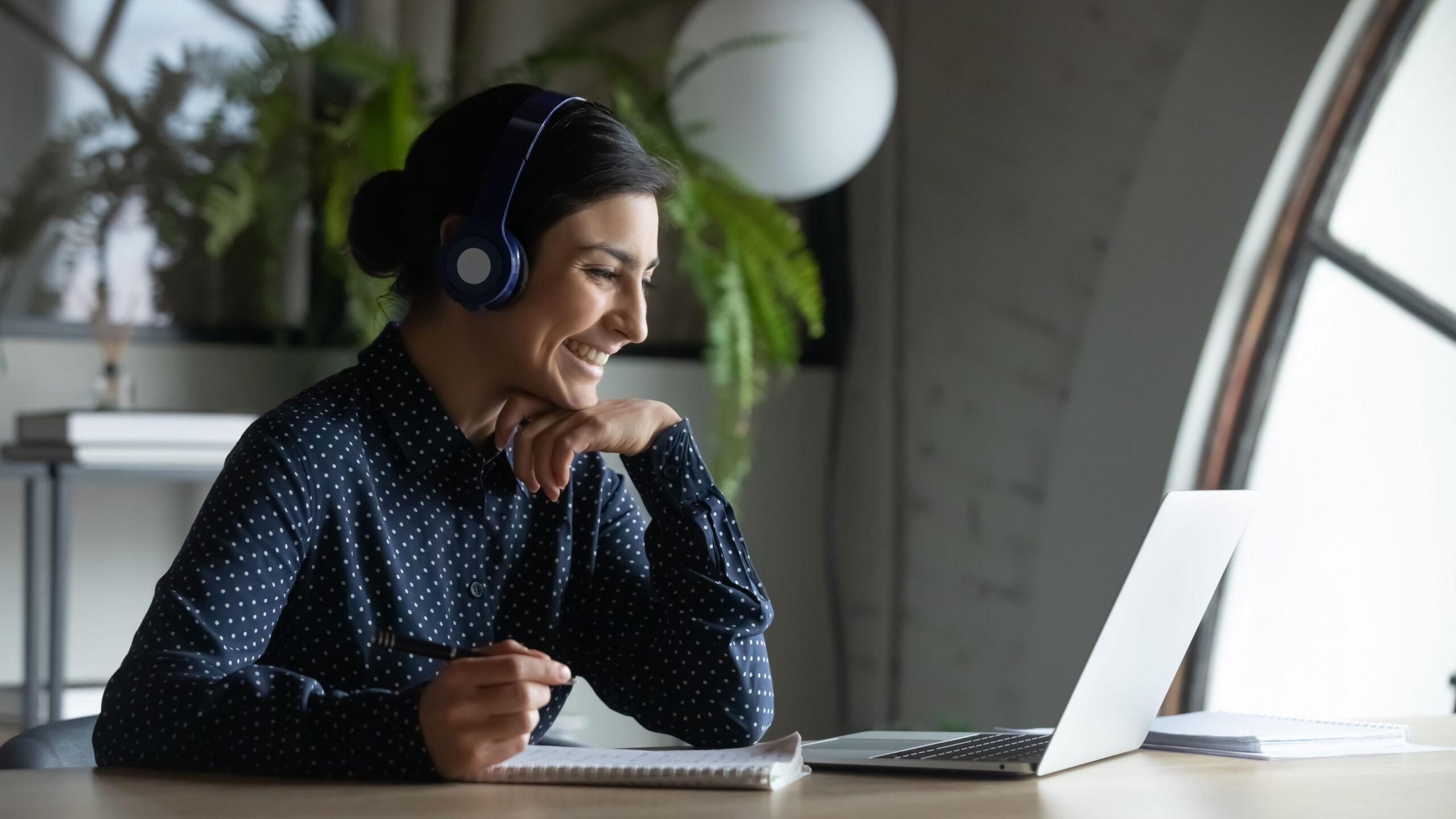 Woman wearing headphones sits at a desk, smiling at a laptop screen with a notebook and pen in hand. Indoor setting with plants in the background.